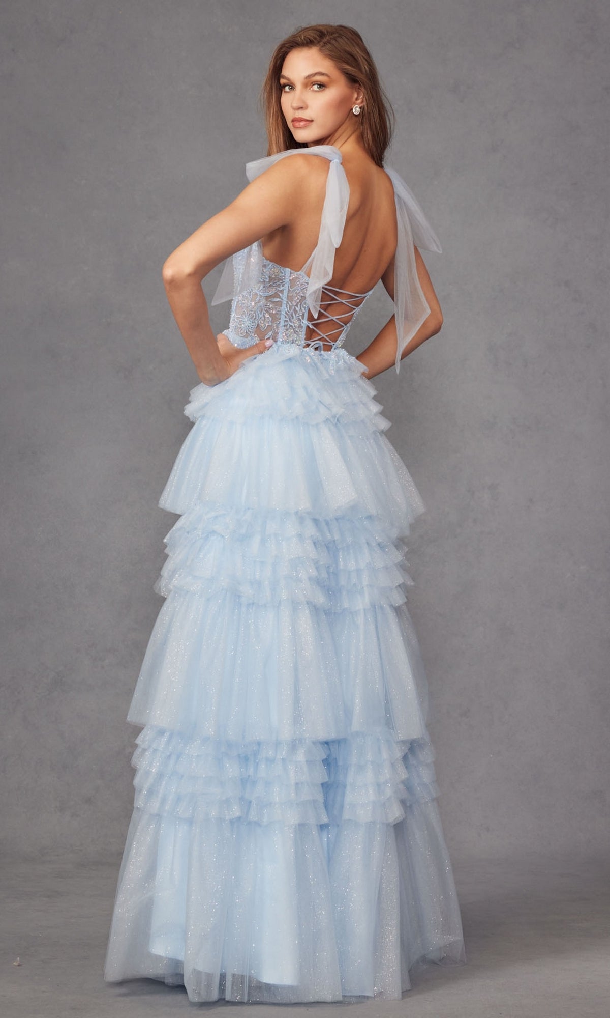 A woman models the Juliet JT2465A Long Ruffled Prom Ball Gown, a light blue tiered tulle dress with sheer ribbon straps, posing against a gray backdrop while looking over her shoulder.